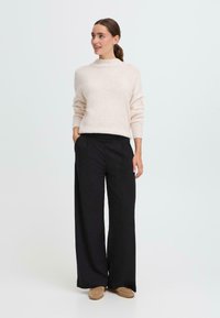 Pull en maille léger beige clair, duveteux, avec un col montant, associé à un pantalon noir à jambes larges. Les chaussures sont des mocassins marron clair. Design simple.