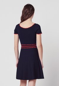 Marindräkt med korta ärmar, vågig röd kant vid hals och ärmar, åtsittande bodice och plisserad kjol. Slät textur, knälång.