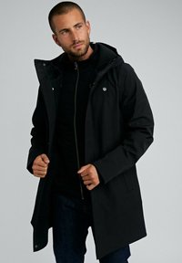 Homme aux cheveux courts et à la barbe ajustant un manteau à capuche noir, vêtu de vêtements sombres et se tenant contre un fond gris clair uni.