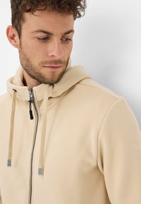 Felpa con cappuccio beige con zip e stringhe, realizzata in tessuto morbido. Presenta una chiusura con zip e dettagli accentuati blu e grigi.