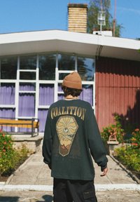 Ciemnozielony, za duży sweatshirt z grafiką na plecach, przedstawiający wzór oka i słowo "KOALICJA". Nosi go z beżową dzianinową czapką.