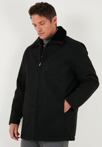 Melns mētelis ar rāvējslēdzēju un kažokādas apdari ap kaklu. Tam ir sānu ieslēgumi un taisna griezuma siluets. Materiāls izskatās maigs un gluds.