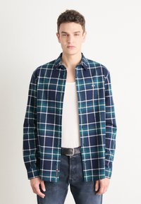 Camisa de franela a cuadros en azul marino y verde azulado, con líneas de rejilla blancas, diseño de botones y un pequeño logotipo en el bolsillo del pecho.