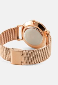 Montre en mailles rose gold avec un boîtier rond, un bracelet en maille et une fermeture à clip. Dos lisse avec des détails minimaux.