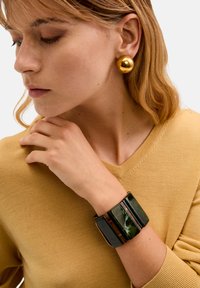 Pulsera gruesa verde y negra con segmentos rectangulares. Aretes esféricos dorados. Lleva un suéter liso color mostaza.