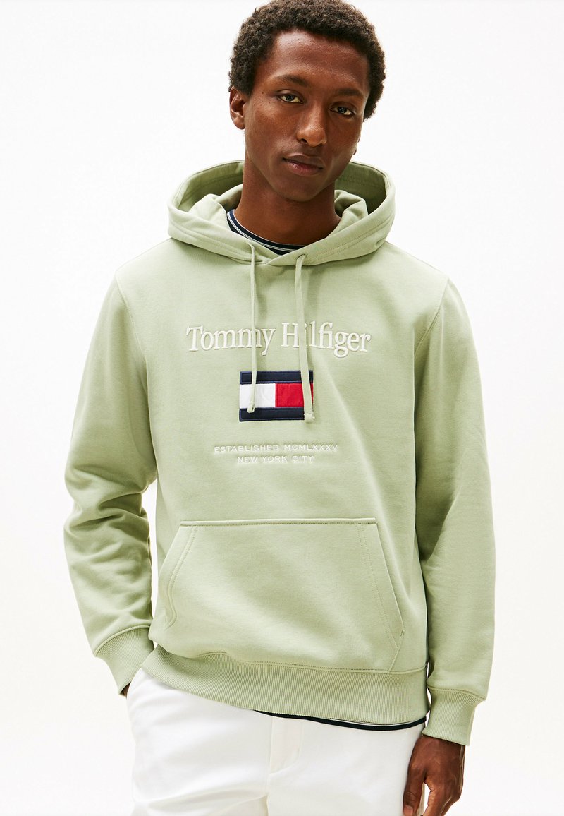 Felpa verde chiaro con tasca a canguro, testo ricamato "Tommy Hilfiger" e logo bandiera. Il materiale sembra morbido; il design presenta polsini a costina.