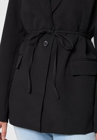Blazer negro de tejido suave, con un cinturón atado en la cintura, cierre de un solo botón y dos bolsillos frontales.