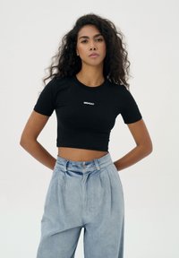 Zwarte cropped t-shirt met korte mouwen, voorzien van het woord "MISAGA" in het wit. Gecombineerd met lichtblauwe high-waisted, wijde spijkerbroeken.