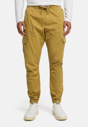 Homme portant un pantalon de jogging moutarde avec poches cargo et des baskets blanches marquées "BACK70", debout les mains détendues le long du corps.