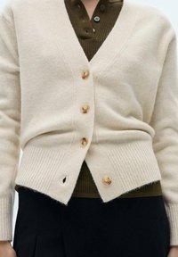 Cardigan en tricot beige avec col en V, boutons en bois, ourlet côtelé et une petite fente latérale, porté sur une chemise à col vert foncé.