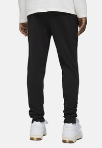 Schwarze Baumwoll-Jogginghosen mit schmal zulaufendem Schnitt, von hinten gezeigt. Kombiniert mit weißen Turnschuhen mit goldenem Logo.