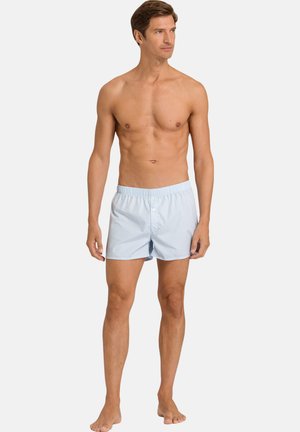 Hemdloser Mann, barfuß stehend, trägt hellblaue karierte Boxershorts, schaut nach links vor einem schlichten weißen Hintergrund.
