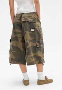 Bermudas cargo camufladas em verde e castanho. Apresenta vários bolsos, um ajuste relaxado e bainhas ajustáveis para personalização. Rótulo da marca exibido.