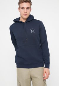 Σκούρο μπλε φούτερ από βαμβάκι με μπροστινή τσέπη, κουκούλα με κορδόνια και κεντημένο λογότυπο "HACKETT H". Απαλή υφή και άνετη εφαρμογή.