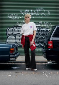 Biała koszulka z kolorowym logo, czerwony sweter związany w talii, czarne spodnie o szerokich nogawkach i srebrne buty z czerwonymi akcentami na tle graffiti.