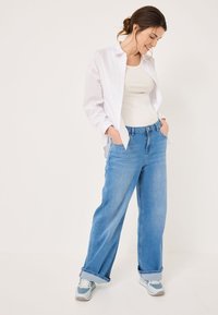 Camicia bianca a maniche lunghe sopra un top beige aderente, abbinata a jeans a gamba larga blu chiaro e scarpe da ginnastica blu con accenti bianchi.