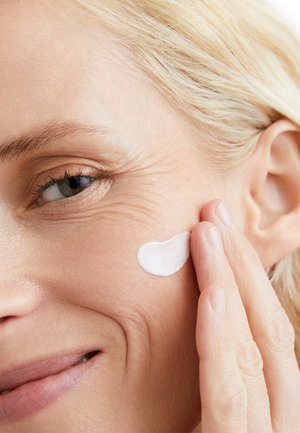 Close-up of a face applying a white cream to the skin. Visible wrinkles, light skin tone, and a slight smile. Background is blurred.