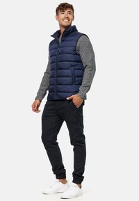 Marineblauer Puffer-Weste mit hohem Kragen, gestepptem Design und grauen langen Ärmeln, kombiniert mit dunklen Hosen und weißen Sneakers.