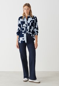 Langärmliges Button-Down-Hemd in Marineblau und Hellblau mit abstrakten Mustern; kombiniert mit dunklen Jeans im Schlag und weißen Turnschuhen.