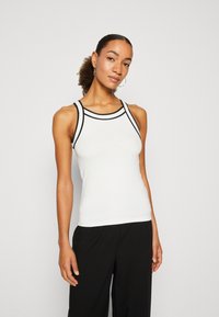 Witte tanktop met zwarte accenten, geribbelde textuur en sportief ontwerp. Beschikt over brede schouderbanden en een aangesloten silhouette. Gecombineerd met zwarte broeken.