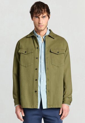 Giacca verde oliva con bottoni, due tasche sul petto, bottoni neri e una texture liscia, indossata sopra una camicia celeste con bottoni.