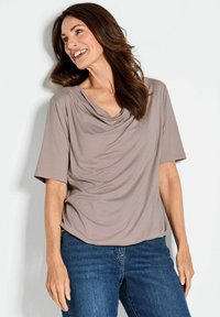 Licht taupe top met col en korte mouwen; zachte, gedrapeerde stof. Gecombineerd met blauwe jeans. Model heeft lang, golvend bruin haar.