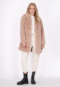 Palton din blană artificială, de culoare bej deschis, cu închidere prin butoane, guler mare și buzunare laterale, asortat cu un top alb și pantaloni crem, cizme negre până la gleznă.