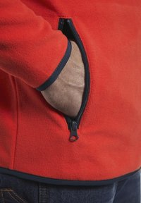 Hand gestoken in de opengewaaide zak van een rode fleecejack, gedragen met donkerblauwe jeans.