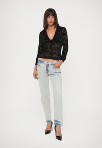 Woman wearing a sheer black lace button-up top, light wash jeans, and black high heels, standing with one hand in pocket.