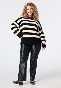 Pull rayé noir et crème à manches larges, associé à un pantalon noir en simili cuir. Chaussures ornées de clous ; cheveux bouclés.