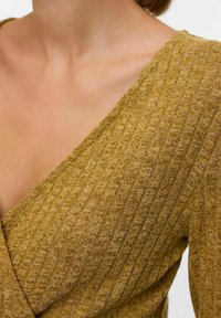 Kollane, tekstureeritud kudum, millel on sügav V-kaelus, diagonaalne ribikiri ja vabalt langev lõige.