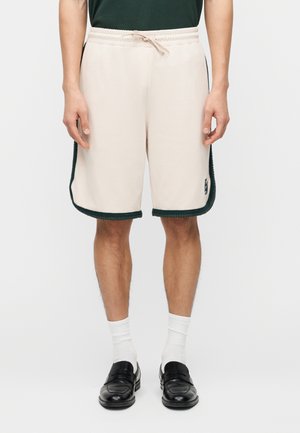 Vyras stovi, vilkėdamas smėlio spalvos šortus su tamsia apdaila, baltas kojines bei juodus odinius batelius, priešais paprastą foną.