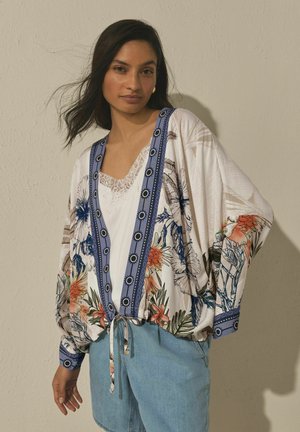 Vrouw met lang donker haar draagt een bloes met kimono-stijl met bloemenprint over een wit shirt met kanten rand en lichtblauwe spijkerbroek, staand tegen een beige muur.