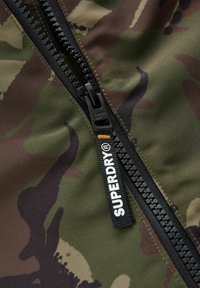 Grüne Camouflage-Jacke mit schwarzem Reißverschluss und Ziehlasche, die mit "SUPERDRY" Branding versehen ist. Der Stoff wirkt glatt und langlebig.