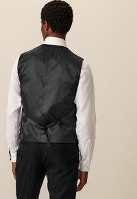 Chaleco de satén negro con una textura suave, detalle de lazo ajustable en la parte trasera, usado sobre una camisa blanca, combinado con pantalones negros.