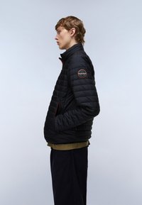 Fiatal személy fekete, steppelt dzsekiben, ujján logóval ellátott folttal, zsebében a kezeit tartva, profilból áll világosszürke háttér előtt.