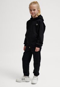 Svart hoodie och sweatpants-set i mjukt material, med en framficka och ett litet emblem på bröstet; kombinerat med vita sneakers.