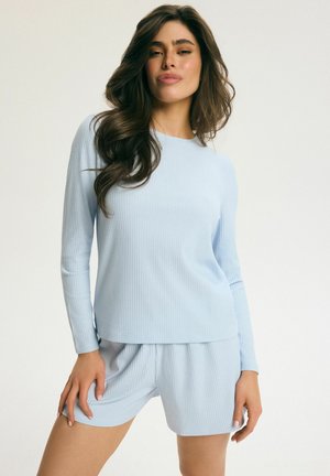 Žena s dlhými vlnitými vlasmi, oblečená v svetlomodrom rebrovanom tričku s dlhým rukávom a zodpovedajúcich kraťasoch, stojaca pred jednoduchým svetlým pozadím.