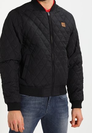 Mand iført en sort quiltet jakke med lynlås og ribkanter ved ærmer og kant, kombineret med blå jeans, mod en hvid baggrund.