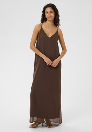 Femme aux cheveux bouclés portant une robe longue marron sans manches avec des fines bretelles et des sandales à talons beiges, debout devant un fond clair.