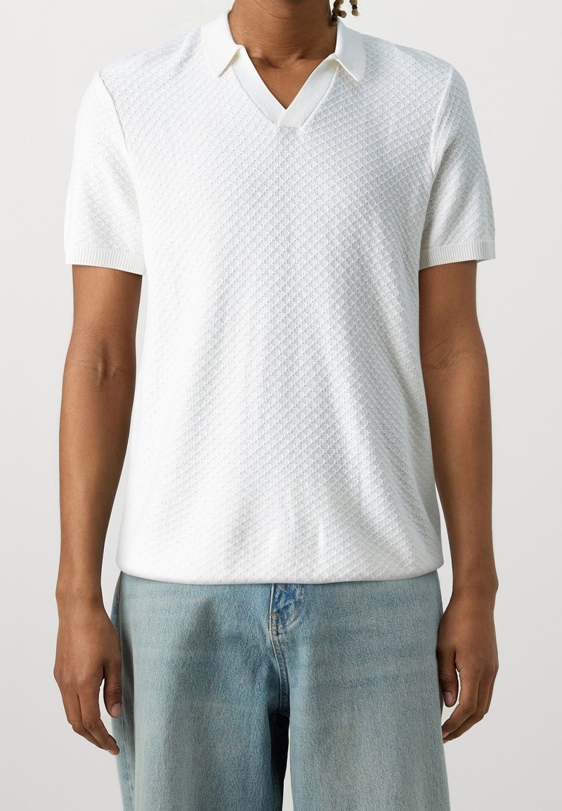 Homme portant un polo blanc texturé à manches courtes et un jean en denim bleu clair, debout devant un fond uni.
