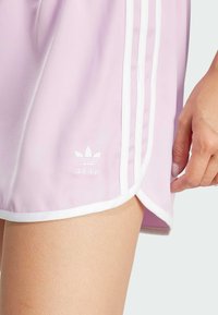 Pantaloni corti sportivi lilla con bordini bianchi, caratterizzati da tre strisce bianche sui lati e un piccolo logo bianco sull'orlo.