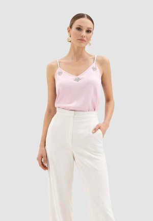 Donna che indossa un top rosa chiaro senza maniche con decorazioni e pantaloni bianchi a vita alta, posa con una mano in tasca.