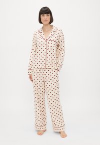 Cremefarvet pyjamasæt med røde prikker, der har lange ærmer, en spids krave og matchende bukser med manchetter og lommer.