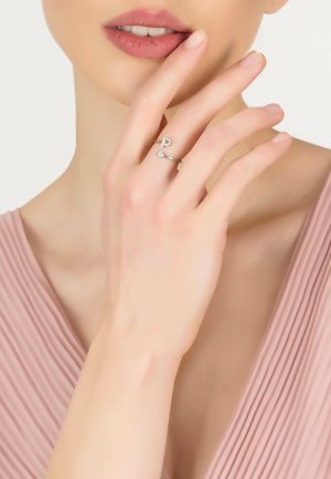 Silver rings with crystal accents on a hand, set against a pale pink background, showcasing a delicate design and minimalist style.