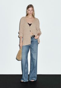 Beige stickad cardigan med en lös passform, knappstängning, bärs över en svart topp, ihop med ljusblå vida jeans och en beige väska.