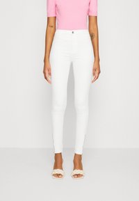 Vaqueros ajustados blancos de tiro alto con detalles de cremallera en el tobillo. Combinados con una blusa ajustada rosa y sandalias trenzadas beige.