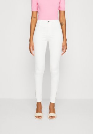 Vaqueros ajustados blancos de tiro alto con detalles de cremallera en el tobillo. Combinados con una blusa ajustada rosa y sandalias trenzadas beige.