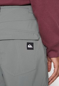 Pelēki šorti ar teksturētu audumu, kuriem ir sānu kabata un melns Quiksilver logo birka. Jostasvieta ir daļēji pārklāta ar bordo krāsas sporta jaku.