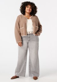 Bruine, textuurrijke fleece jas met witte bies; drie knopen en twee voorzakken. Gecombineerd met lichtgrijze wijde jeans en schoenen met luipaardprint.